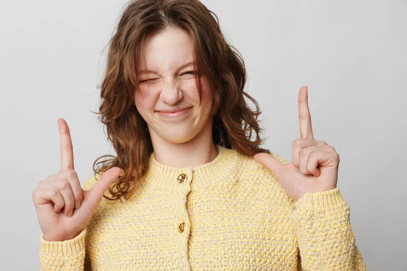 Young woman in yellow sweater makes finger gun gesture with closed eyes and playful smile. Studio portrait on grey background. Concept of fun, youth, and casual expression.