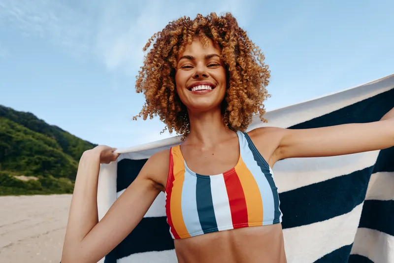 Happy young african american woman with curly hair enjoys sunny day on the beach wearing a colorful striped swimsuit top and holding a towel behind her shoulders.