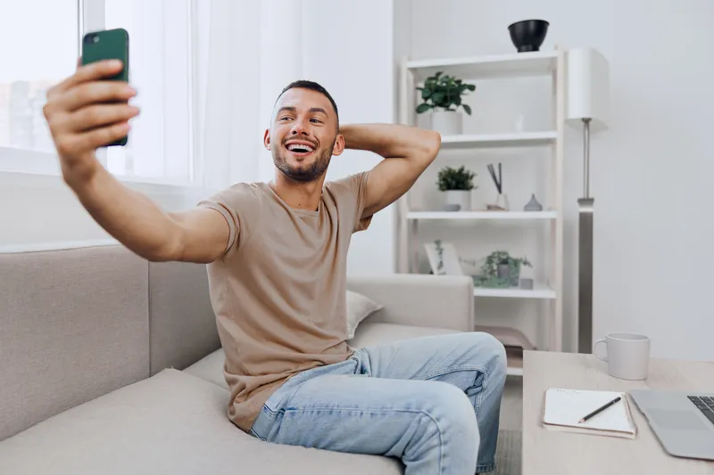 Engaging man smiling while taking a selfie at home, showcasing genuine joy and soft masculinity in a cozy living space filled with plants.