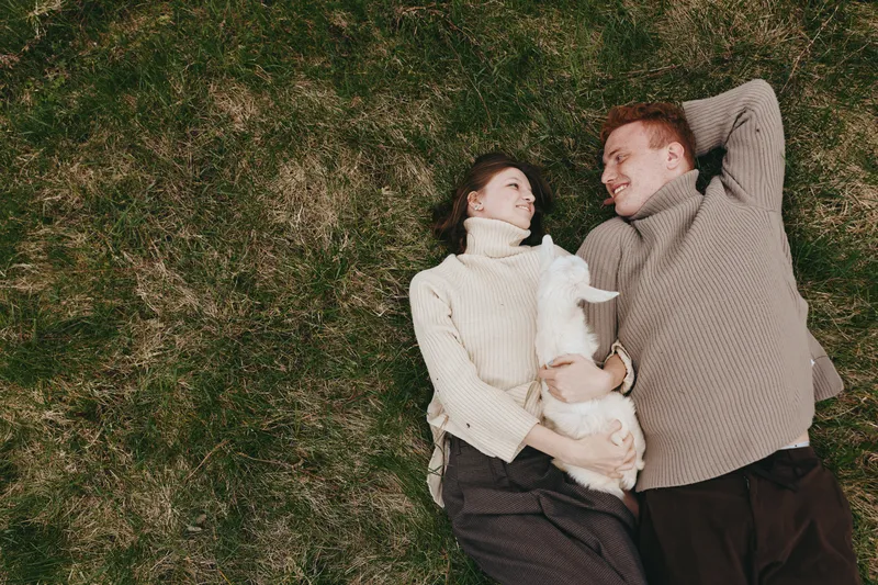 A relaxed couple lies on green grass with a fluffy dog between them, smiling and cuddling under soft sweaters, enjoying a sunny outdoor moment. Concept for companionship, affection, and everyday