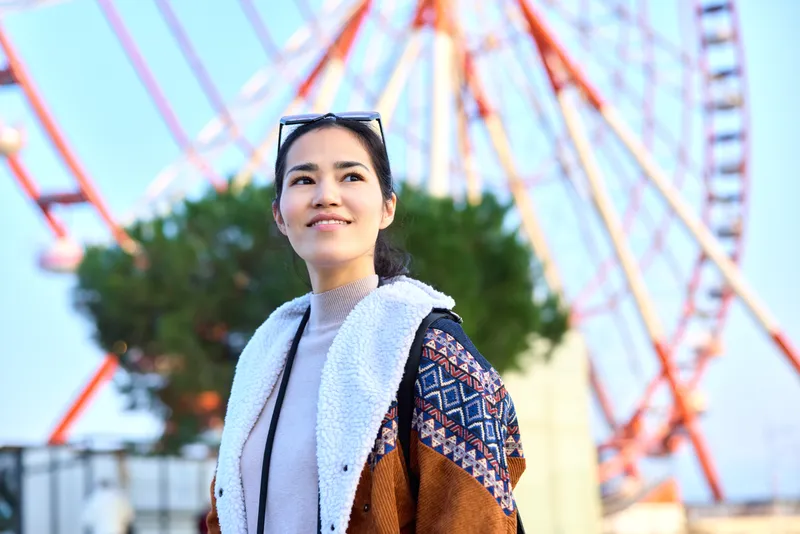 Woman outdoors with scarf and jacket beside a ferris wheel, sunny mood, urban style, and a confident smile, capturing a fashionable travel moment in a vibrant cityscape today