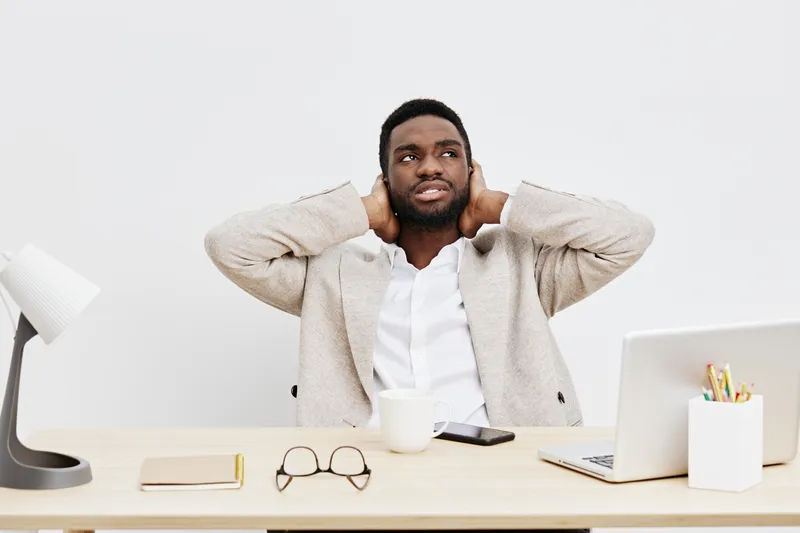 Professional young man in casual clothing seated at desk in office workspace, smiling, relaxed, with a laptop, glasses, cup, and stationery on white background, business and remote work concept.
