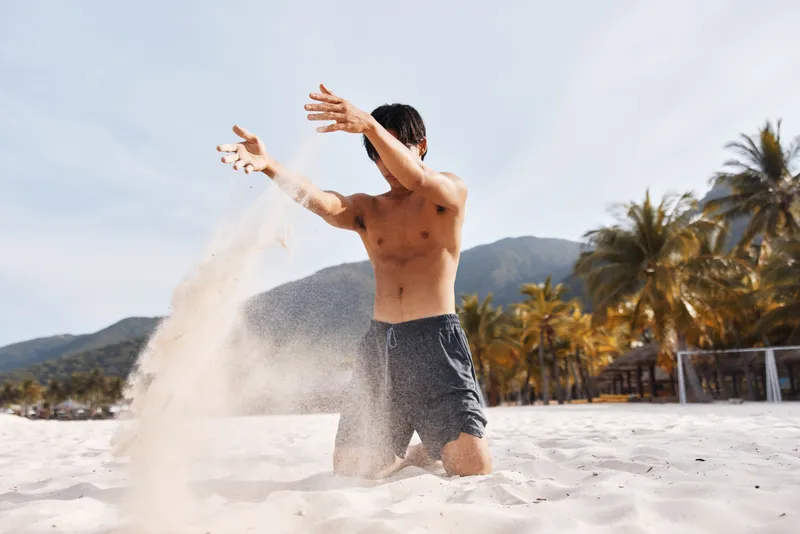 Мускулистый азиатский мужчина наслаждается отдыхом на тропическом пляже, счастливо улыбаясь и демонстрируя обнаженный торс