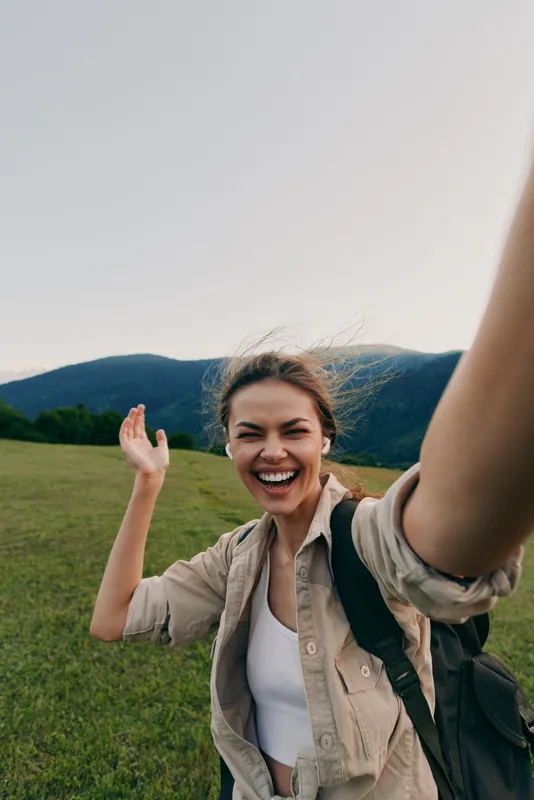 Happy young woman takes selfie outdoors in green field with mountains in background, wearing casual clothes and backpack, enjoying nature and lifestyle.