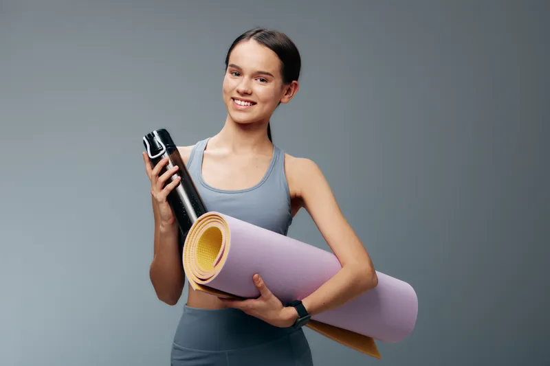 Fitness woman in athletic wear holds a yoga mat and water bottle, ready for a workout. Studio setting highlights sport, training, health, and balance in a energetic routine.