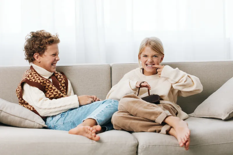 Two children sit on a cozy sofa, smiling and sharing a playful moment. One boy wears a knitted sweater and vest, the other girl holds a soft toy bag and laughs.