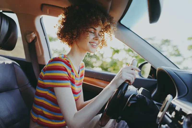 Woman driving a car with a striped shirt and curly hair, enjoying a sunny day, candid lifestyle shot with retro film color, bright interior, cheerful mood