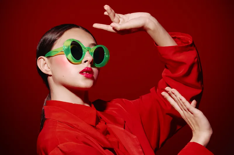 Fashion woman wearing large green round sunglasses and red shirt poses creatively against red background. Bright makeup and dramatic hand gesture create bold beauty concept.