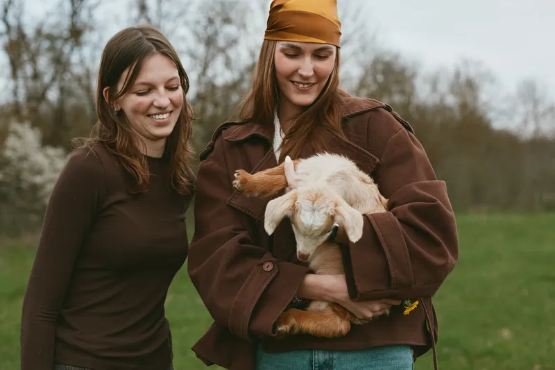 Goat kid held by one woman while another smiles outdoors, warm jackets in a countryside setting, playful moment, wholesome interaction, vibrant natural tones