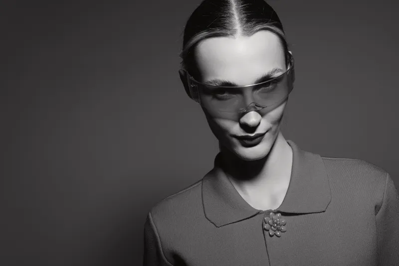 Black and white portrait of a young Caucasian woman with light skin wearing transparent sunglasses and a textured blouse. Studio lighting creates a dramatic mood.