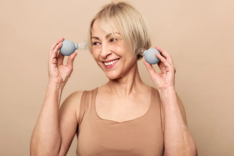 Smiling mature woman with short blonde hair holds two blue facial suction cups near her cheeks. Beige background and natural light highlight skincare, beauty routine, and self-care concept.