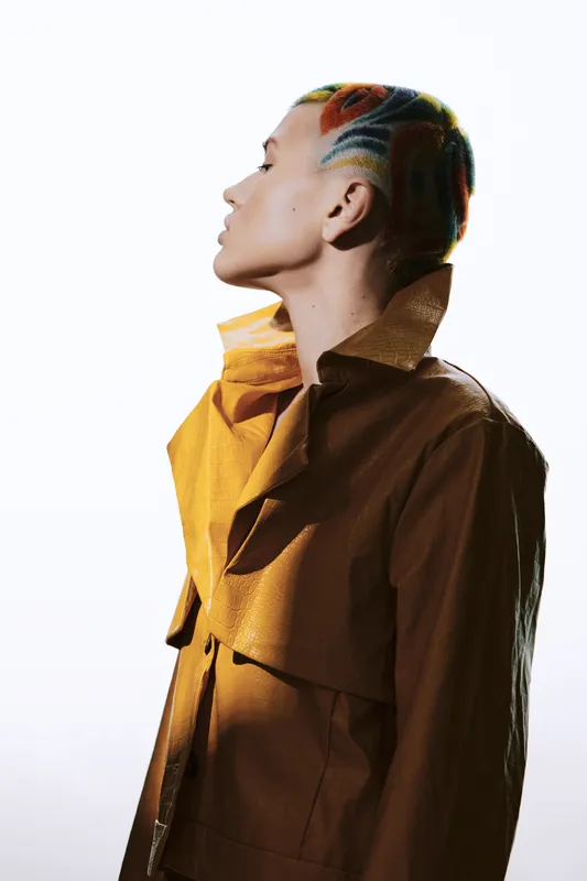 Profile of a woman with colorful short hair in a stylish brown jacket, showcasing modern fashion and artistic expression with a plain white background.
