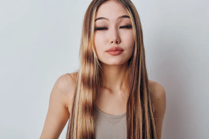 Woman with long straight hair and closed eyes wearing a beige tank top on light gray background. Female portrait with natural makeup and serious expression for beauty concept.
