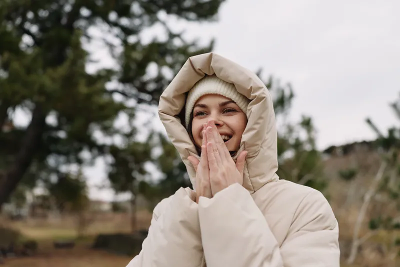 Woman in a beige winter coat outdoors, smiling with hands near her face, cozy portrait in chilly weather and natural surroundings