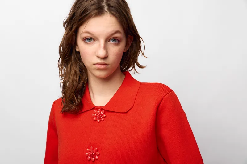 Young woman with wet hair wearing a red textured sweater with floral buttons looks serious on a white background. Studio portrait with natural expression and casual style concept.