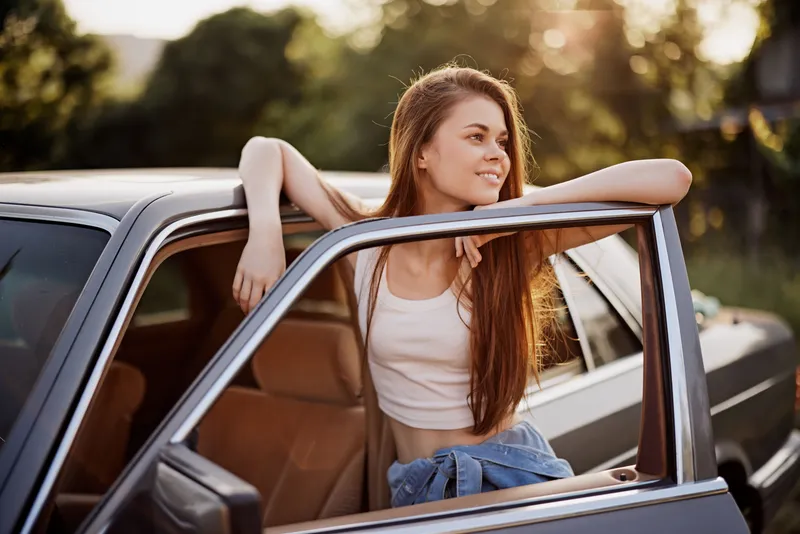 Young woman enjoying a sunny day, leaning out of a vintage car, embodying freedom and adventure with a stylish casual outfit.