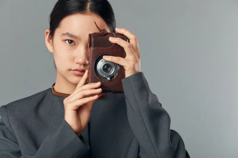 Young woman holding vintage camera close to face looking at viewer against plain gray background wearing dark jacket with minimal makeup and neat hairstyle indoors.