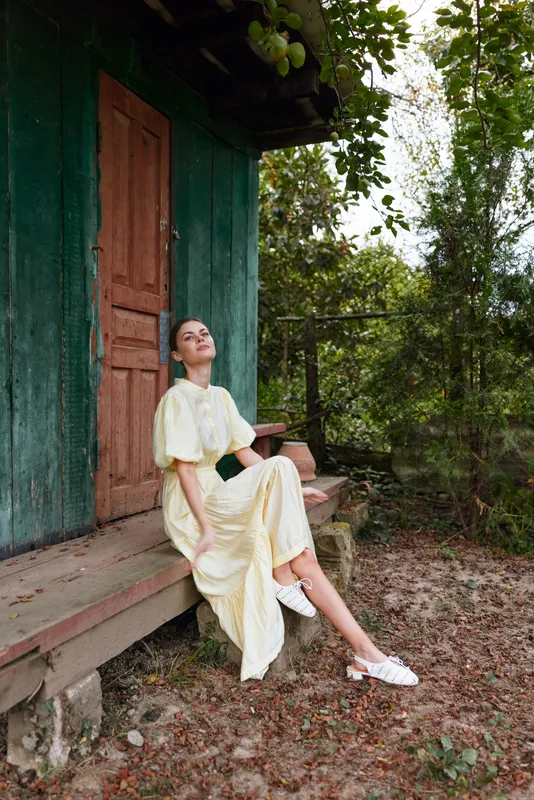 woman sitting outdoors on a rustic porch in a pale yellow dress, serene pose under sunlight, wooden doorway with greenery framing a peaceful natural setting