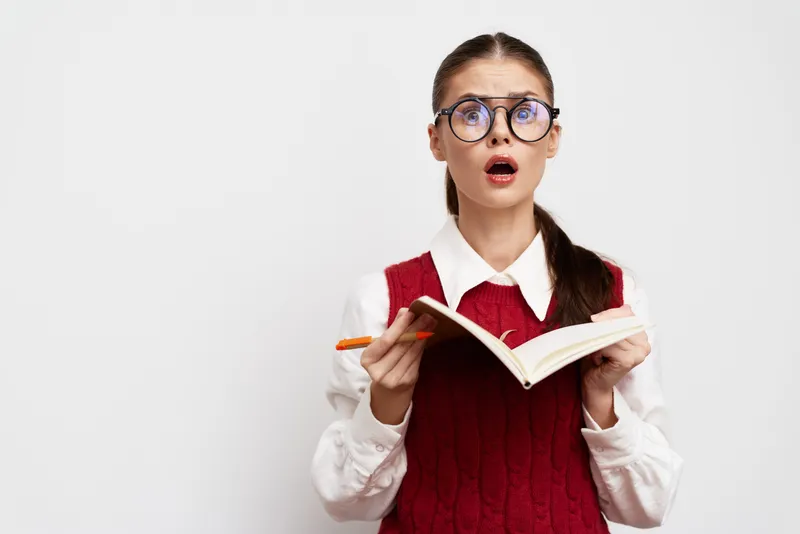 Удивлённая девушка-студентка в очках держит книгу, выглядя потрясённой во время чтения