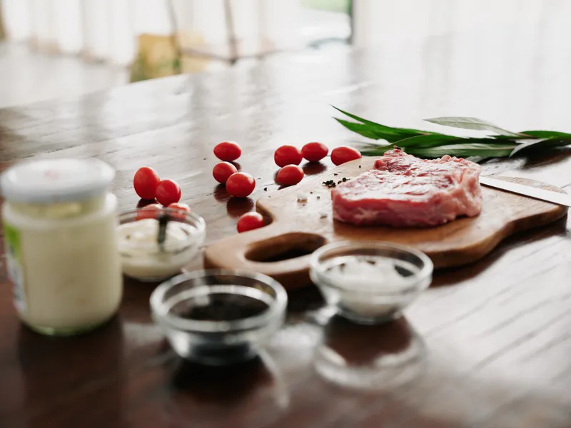 Стильная кухня: на деревянном столе лежит кусок свежего мяса со специями, помидорами и зеленью, из окна пробивается свет от огня