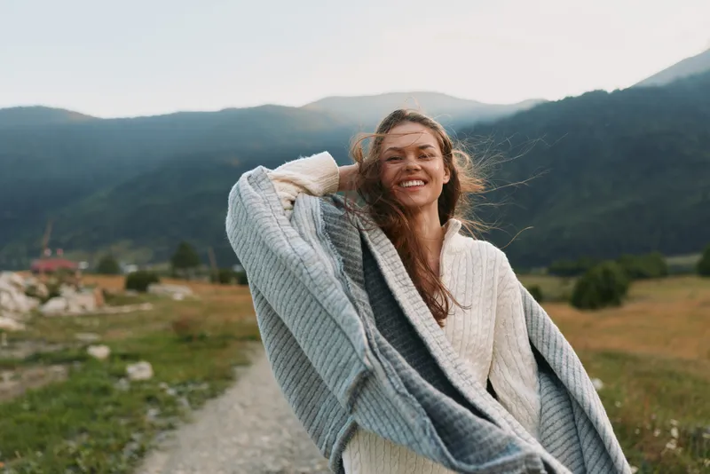 A happy woman in a white sweater and gray shawl smiles while standing on a rural path with mountains in the background. Lifestyle concept of freedom and nature.