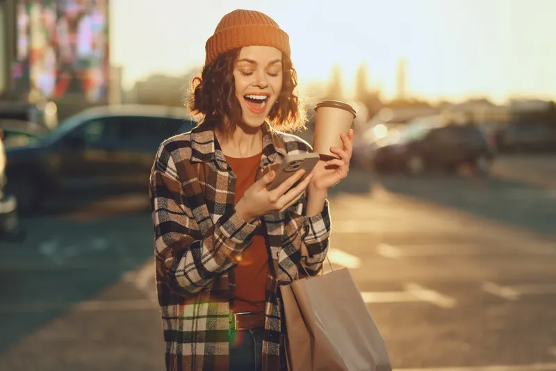 Woman with coffee and phone, smiling while shopping in urban sunlight at golden hour glow. Candid lifestyle portrait with authenticity, mindful living and emotional storytelling mood.