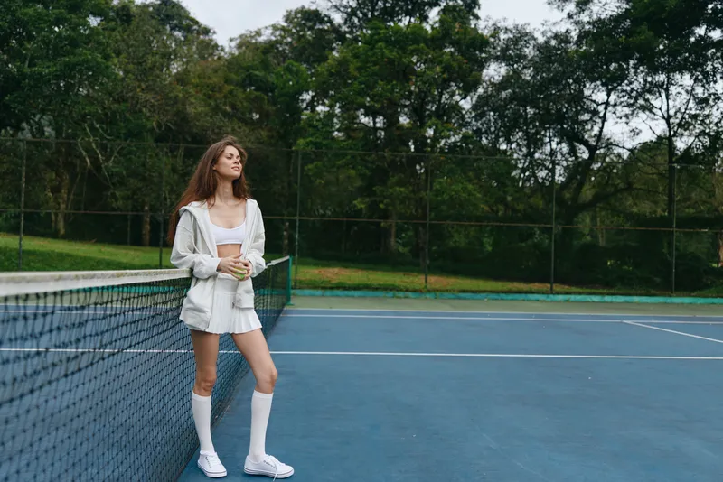 Young female tennis player in sporty outfit enjoying a moment of relaxation on the court surrounded by greenery, reflecting passion for the sport and nature