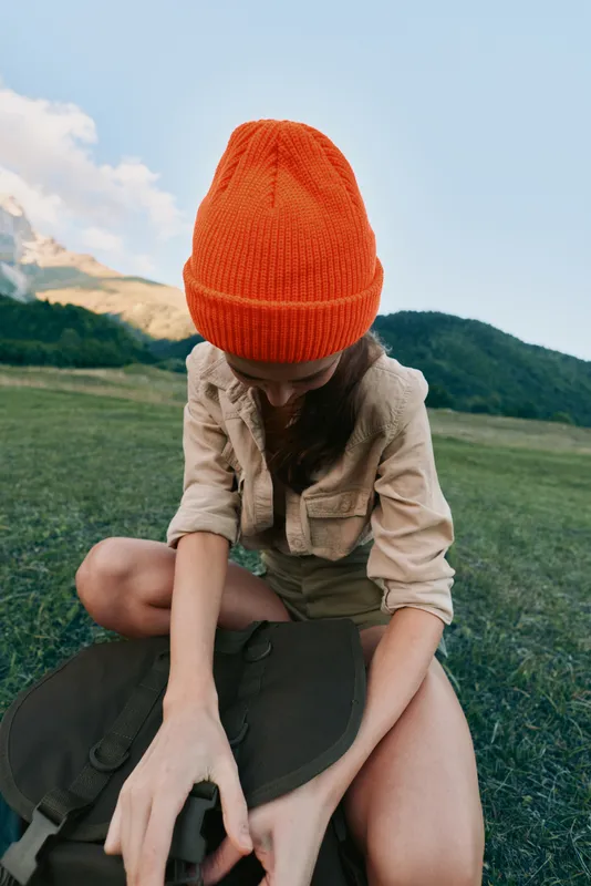 Portrait of a woman in an orange beanie and casual outfit in a grassy field, capturing a relaxed lifestyle moment outdoors. Natural light creates a warm, calm mood.