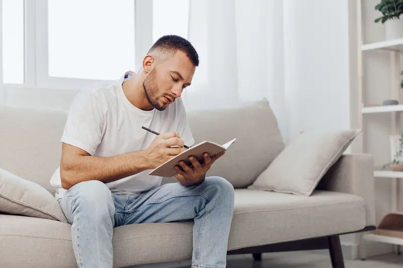 Thoughtful man writing in a notebook at home, wearing casual clothes, surrounded by natural light, reflecting on ideas and creativity. Concept: mindfulness.