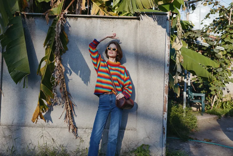 Woman in her mid 20s wearing a colorful striped sweater, jeans and sunglasses leaning against a textured wall with tropical leaves, smiling in a casual crossbody bag lifestyle travel shot.