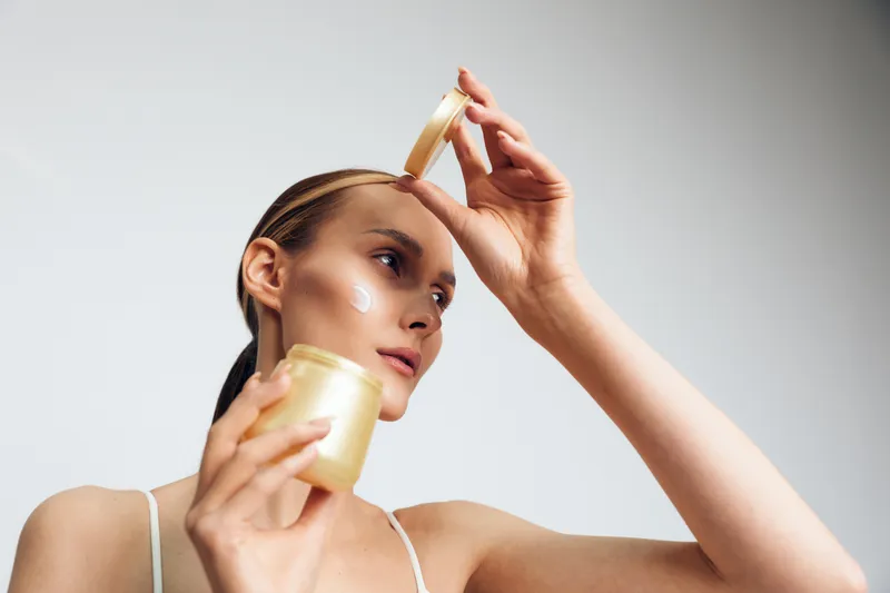 Young woman with slicked back hair applies moisturizer to her face from a jar in a bright minimalist setting, demonstrating skincare and self-care routine.