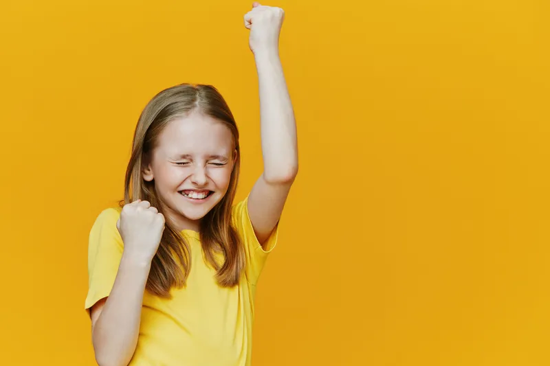 Happy girl celebrating success with raised fists against a vibrant yellow background, wearing a bright yellow t shirt, conveying joy and positivity