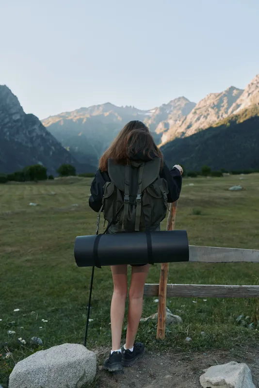 A lone hiker stands in a grassy valley, facing distant mountains. She wears a backpack and boots, ready for a trek in a tranquil, open landscape.