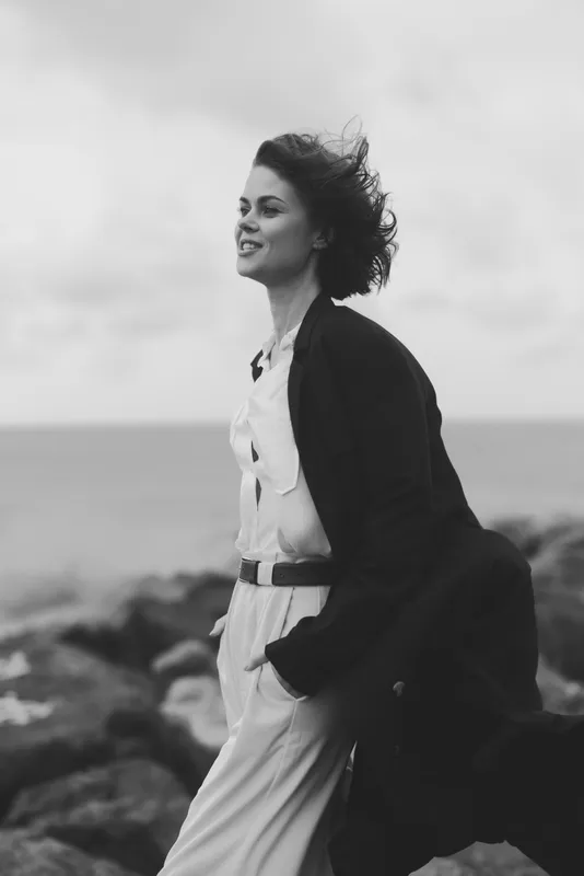 Woman strolling along a rocky shoreline in a flowing dress and overcoat, wind in her hair, black and white scene capturing fashion, movement and coastal mood