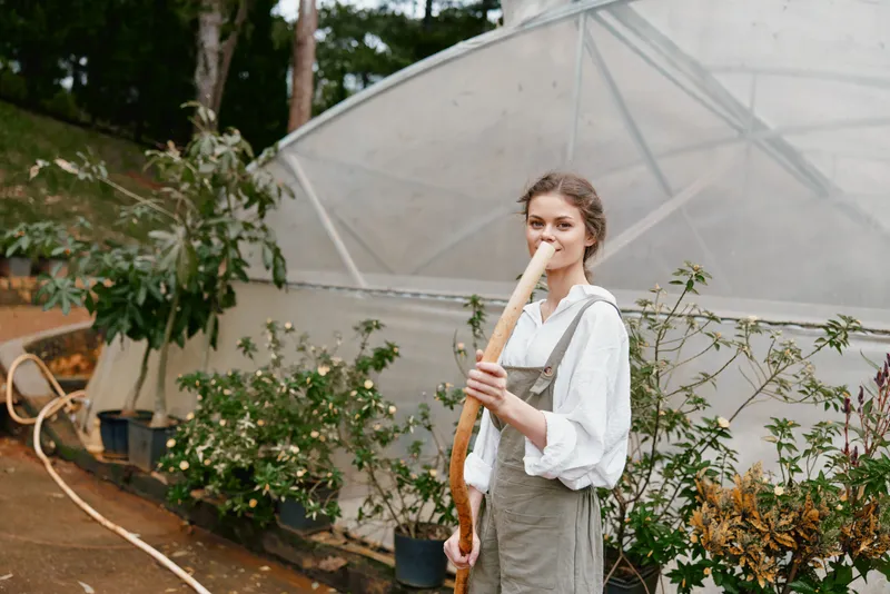 Young woman wearing rustic clothing in a greenhouse, holding a gardening tool, surrounded by plants and greenery for sustainable agriculture
