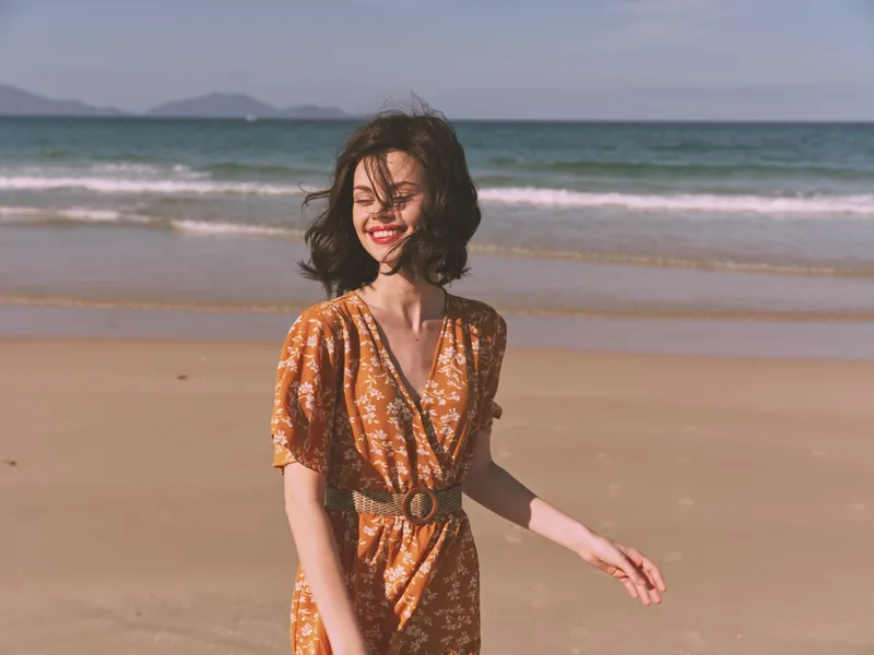 Счастливая женщина в оранжевом платье стоит на песчаном пляже на закате, подняв руки вверх