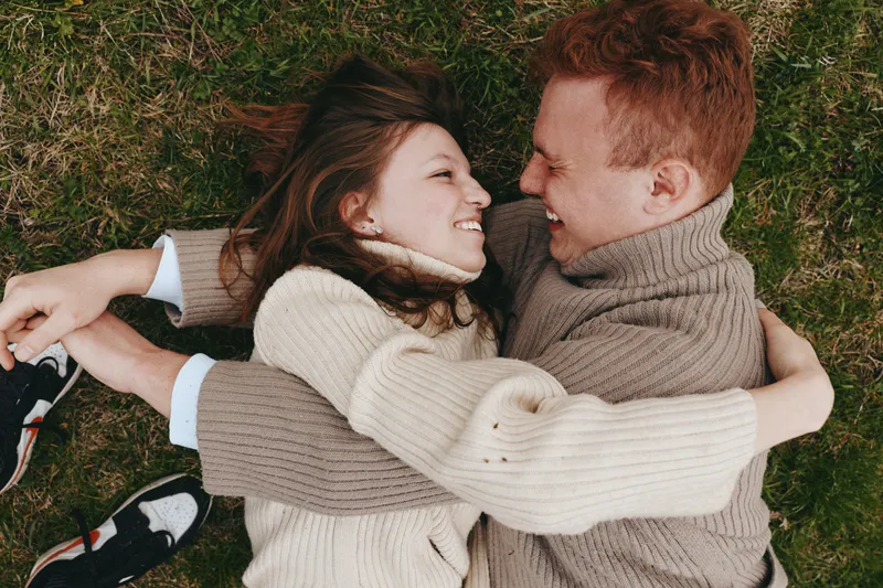 Smiling couple lying on grass embracing outdoors, cozy sweaters, warm light highlighting their connection. Concept of togetherness, affection, and carefree lifestyle