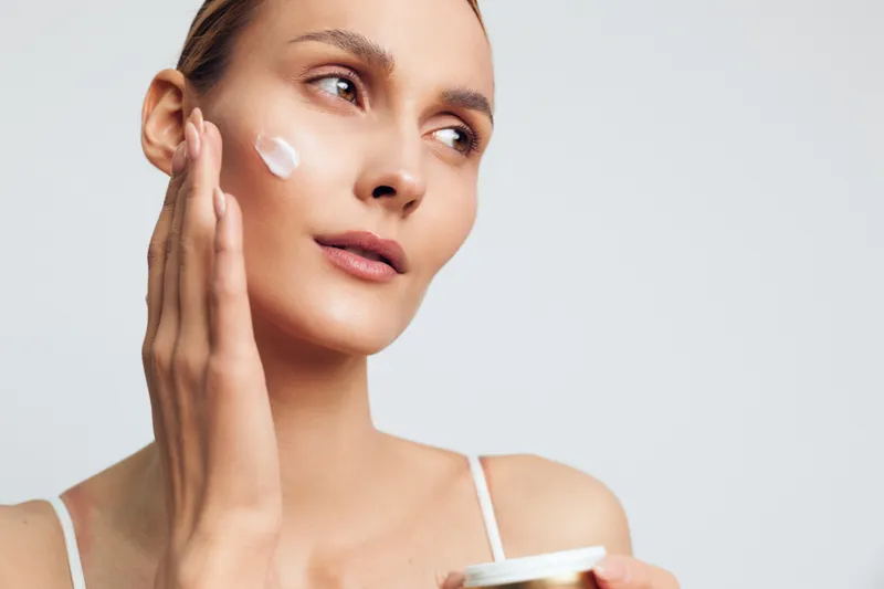 Young woman applies cream to face with gentle touch looking away with clean fresh skin and natural makeup isolated on white background in skincare beauty routine concept.