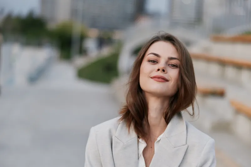 Confident businesswoman smiling outdoors, dressed in a light blazer, with a blurred urban background suggesting professionalism and ambition.