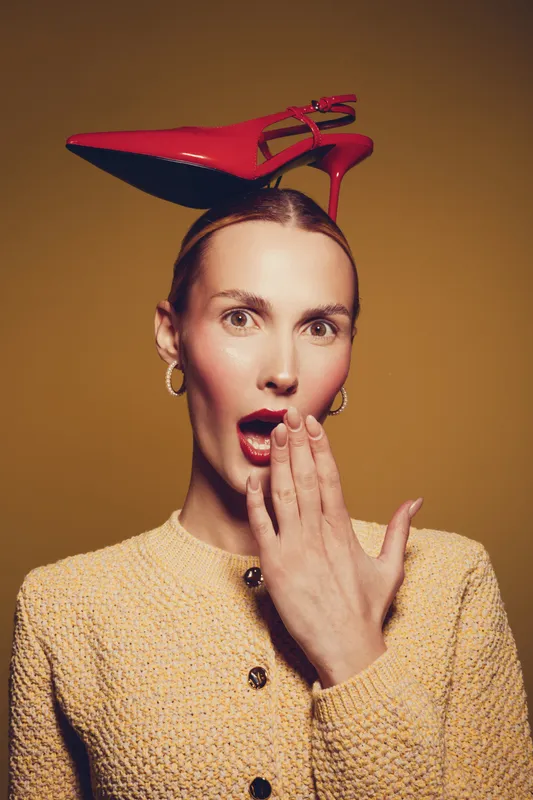 Surprised woman with red high heel shoe balanced on her head, wearing yellow textured jacket and gold earrings. Concept of fashion, style, and playful expression in studio.