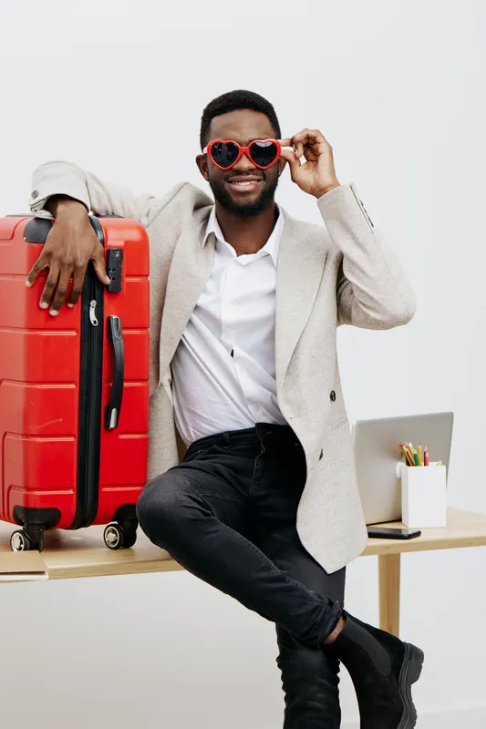 Stylish young man with dark skin smiling and wearing red heart-shaped sunglasses, sitting on desk with a red suitcase and laptop, celebrating travel or vacation plans in a bright minimal studio backgr