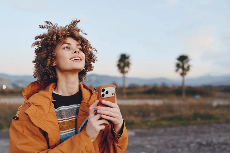 Woman with curly hair holding smartphone outdoors, wearing orange jacket, portrait lifestyle moment, looking up at the sky, soft daylight over open landscape, distant hills, calm happy mood