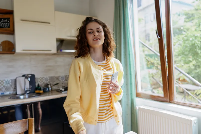 Young woman with curly hair stands in a sunlit kitchen, smiling softly while holding a tablet, a casual home lifestyle moment.