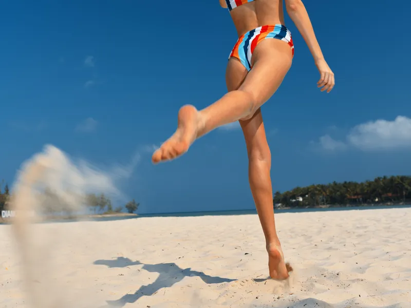 Dynamic beach scene featuring a sunlit figure kicking up sand in a playful stride, wearing a striped swimsuit. Clear blue sky, calm sea, and soft shoreline create vibrant summer energy.