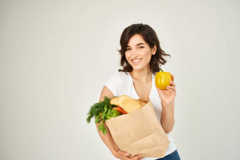 Симпатичная женщина в белой футболке держит продуктовый пакет с овощами, пропагандируя здоровое питание