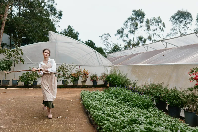 Young woman walking in a greenhouse, wearing an apron and carrying a stick, surrounded by vibrant greenery, reflecting a connection to nature and sustainable living