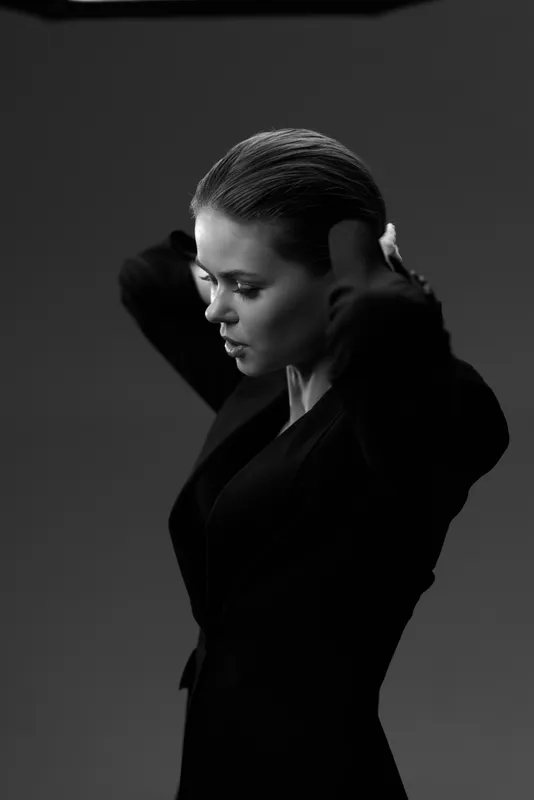 Black and white portrait of a woman adjusting hair in a dark outfit with dramatic lighting and serious expression