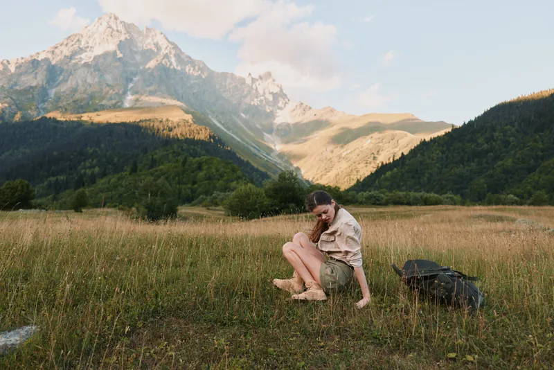 A woman in casual clothes sits on grass in a mountain valley, surrounded by nature and wildflowers, enjoying a peaceful moment in the outdoors lifestyle.