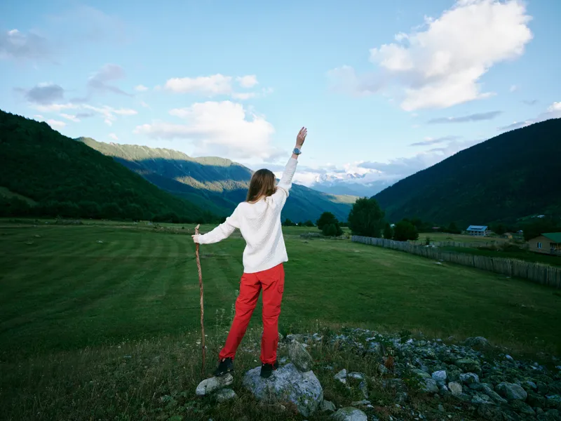 Woman standing on a grassy hillside with a walking stick, raising her arm in triumph as mountains loom in the distance and a wide valley opens to blue sky and lush countryside