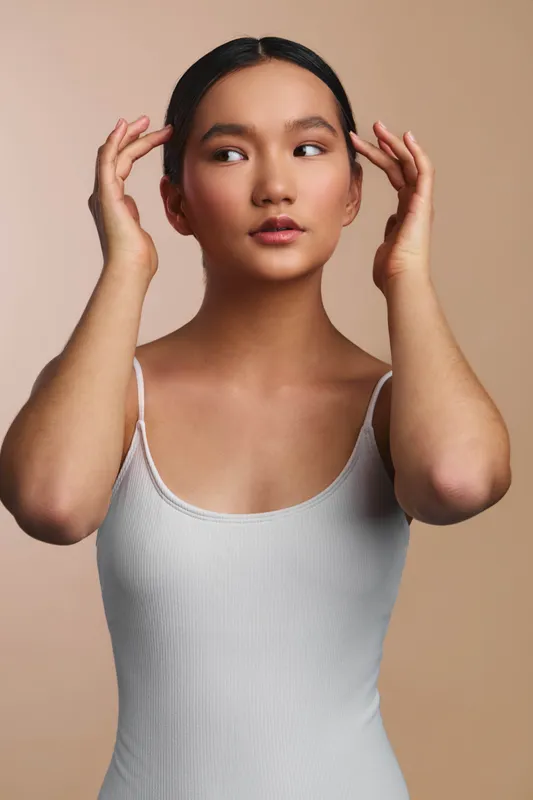 asian woman in a white tank top adjusts her hair, posed against a soft beige backdrop. she gazes slightly off camera, conveying calm confidence and natural poise.